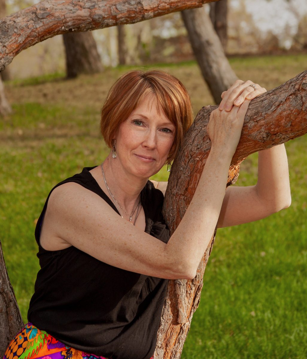 Chris MacPherson resting her arms on a large old tree branch, smiling at the camera